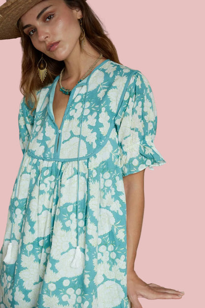 Woman wearing a teal floral dress standing indoors with plants in the background