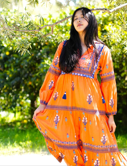 Woman wearing an orange dress with floral patterns standing outdoors among greenery.