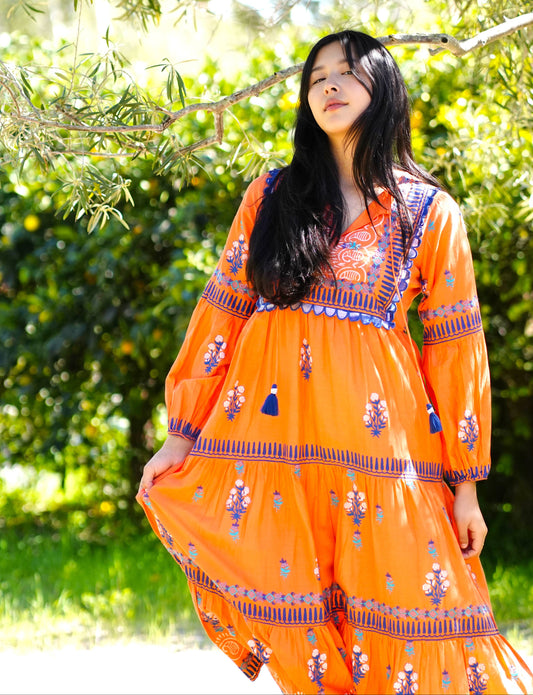 Woman wearing an orange dress with floral patterns standing outdoors among greenery.