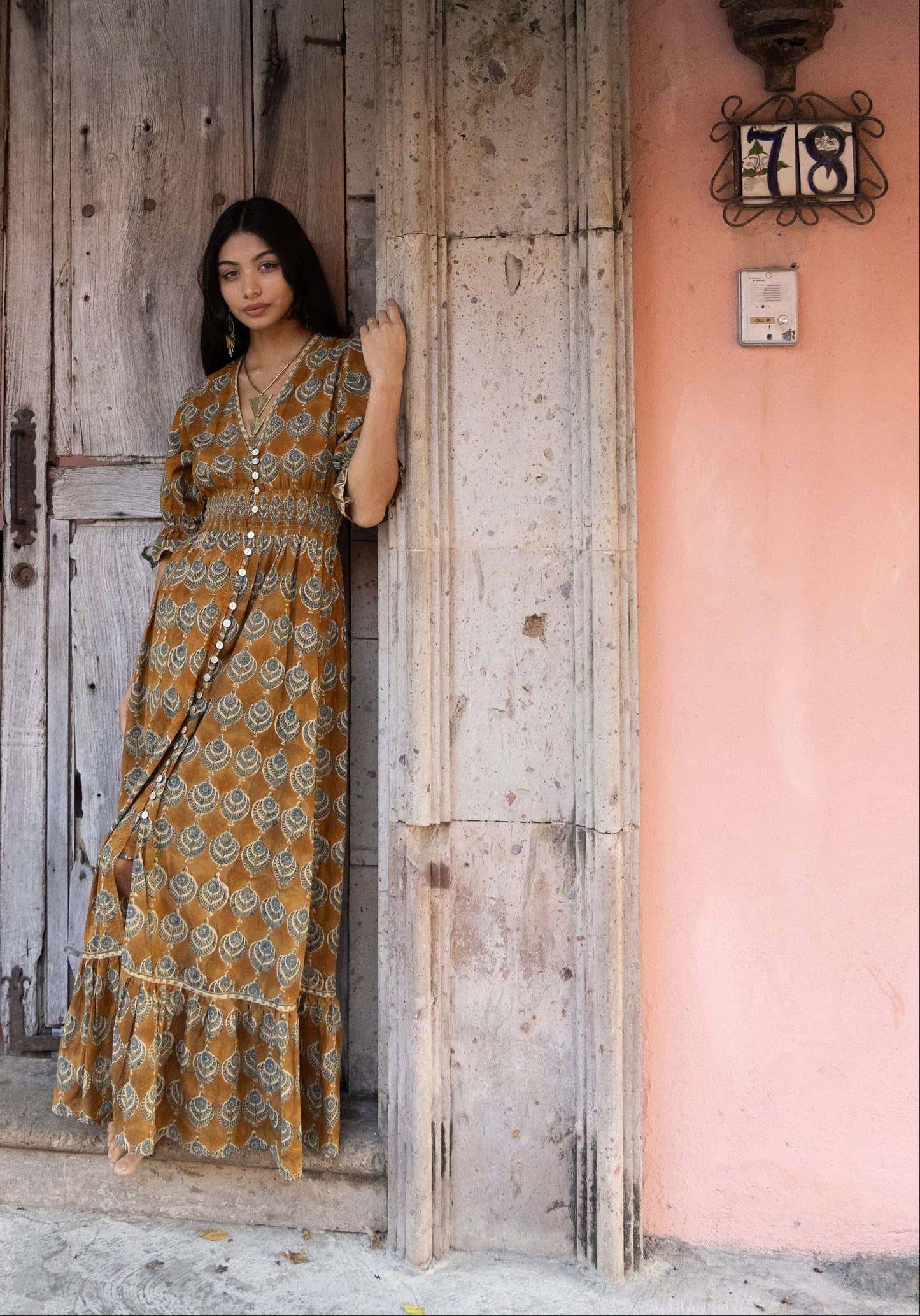 Woman in a patterned dress standing in front of a rustic wooden door with pink flowers above.