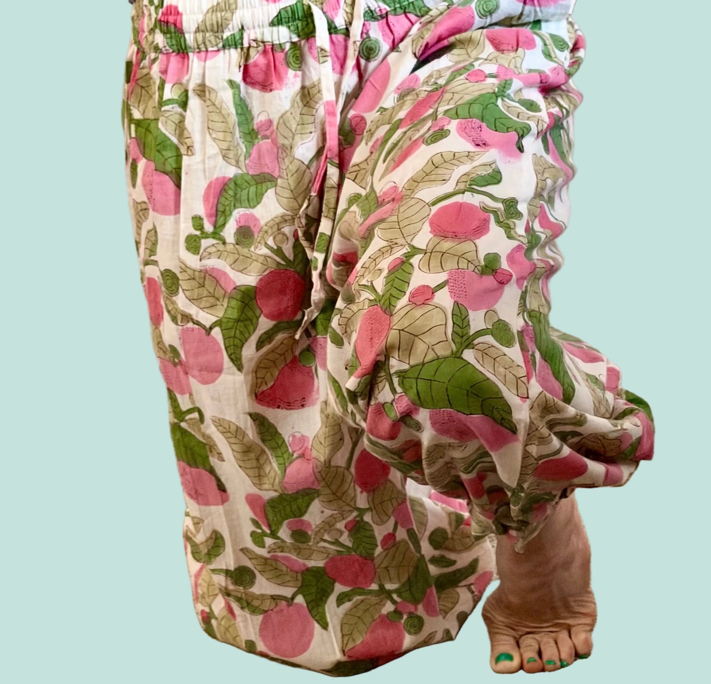 A person wearing loose, gathered pants with a pink and green botanical print on a cream background, standing barefoot on a beige carpeted floor.