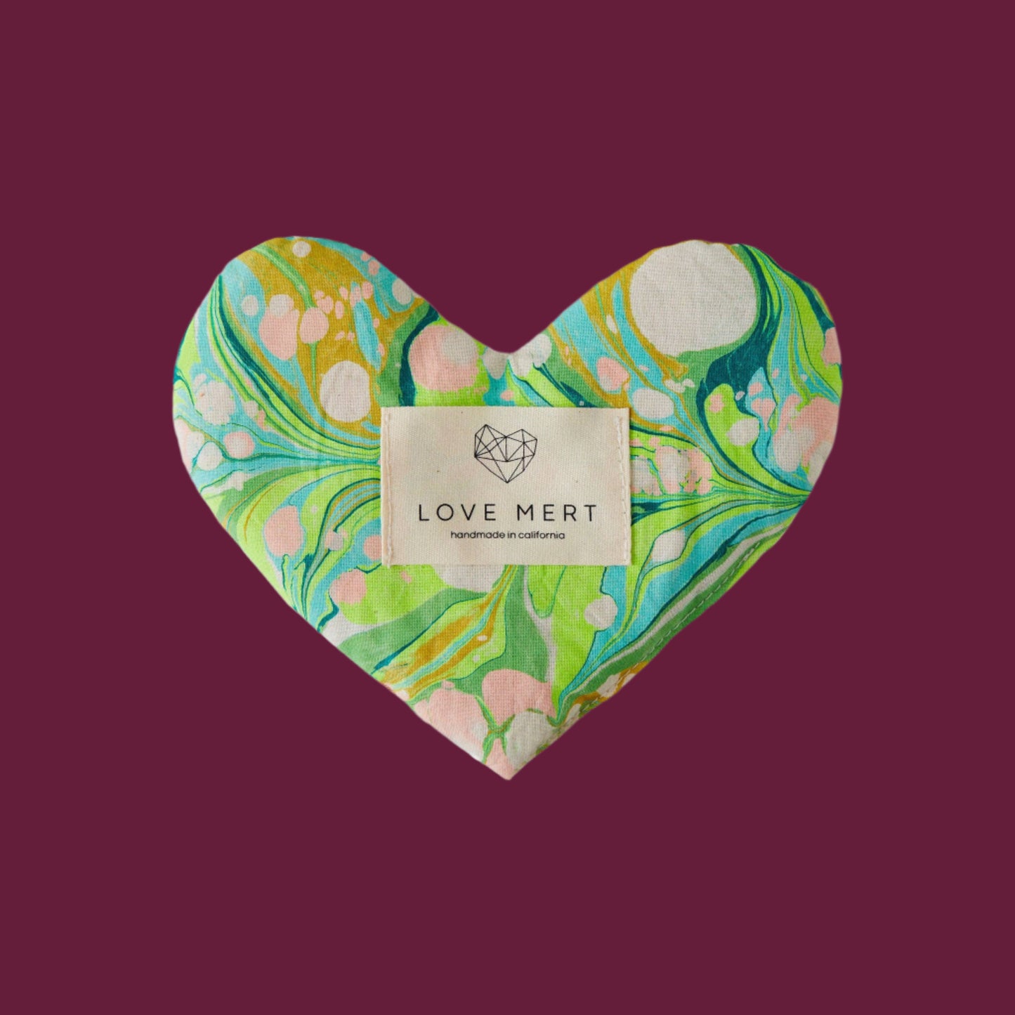 A heart-shaped eye pillow with a marbled pattern of greens, blues, and pinks, featuring a raw silk backing and a label with 'LOVE MERT' branding.