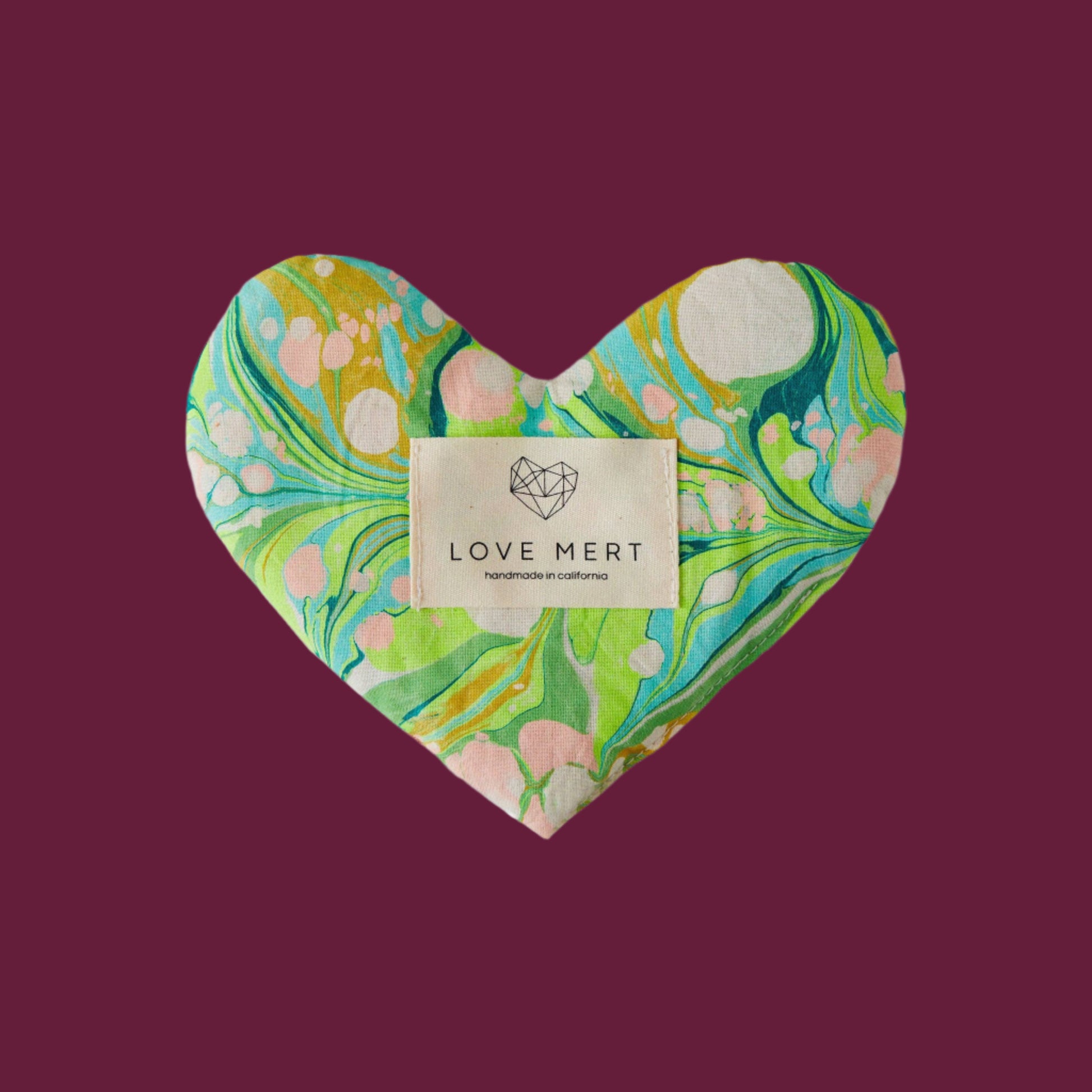A heart-shaped eye pillow with a marbled pattern of greens, blues, and pinks, featuring a raw silk backing and a label with 'LOVE MERT' branding.