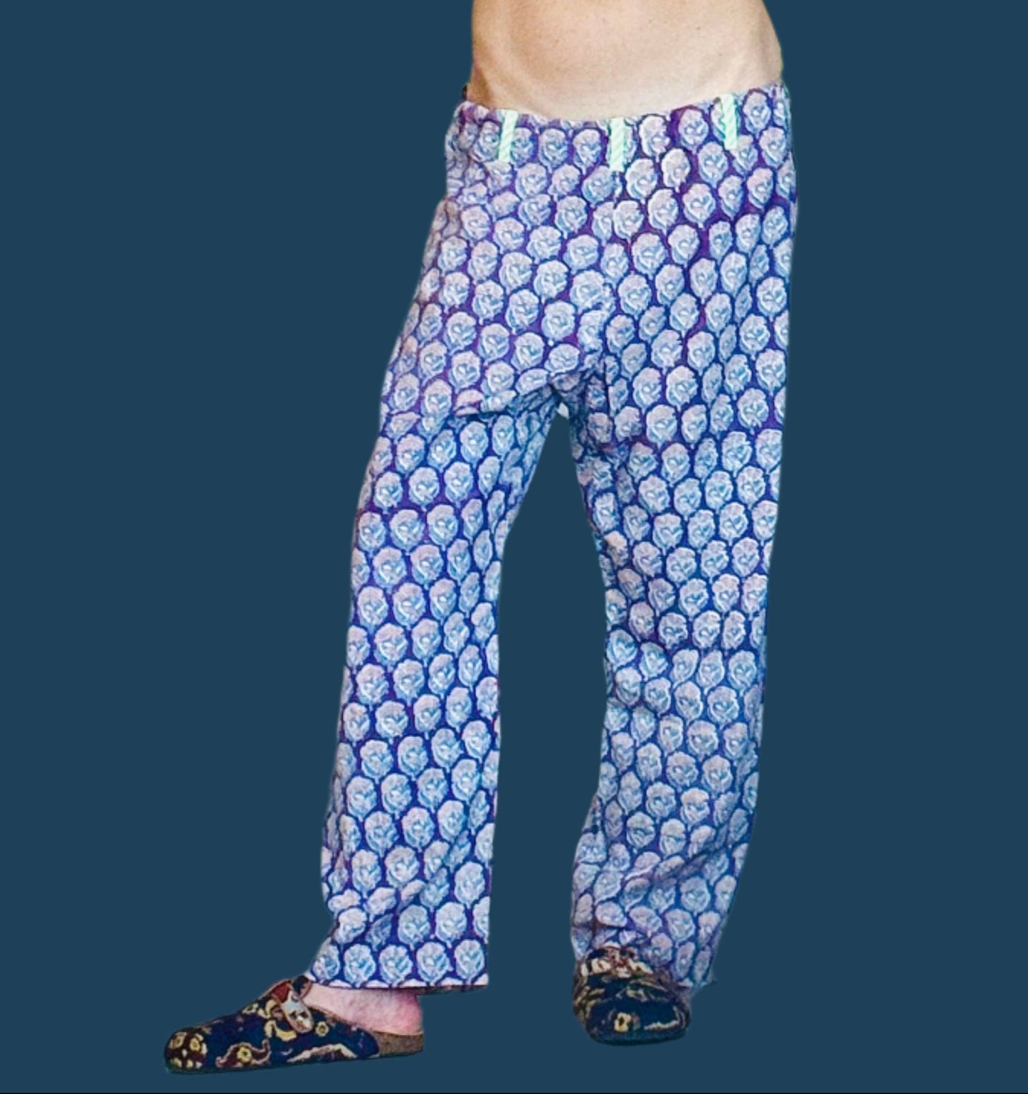 Person taking a mirror selfie wearing a blue top and patterned pants in a room with shelves and clothes.