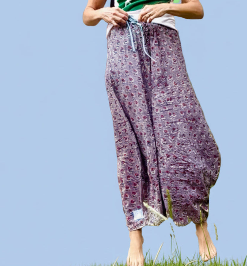 A woman stands outdoors in front of a stone wall and window, wearing loose purple patterned pants and a green-and-white top, with a black crossbody bag across her chest.