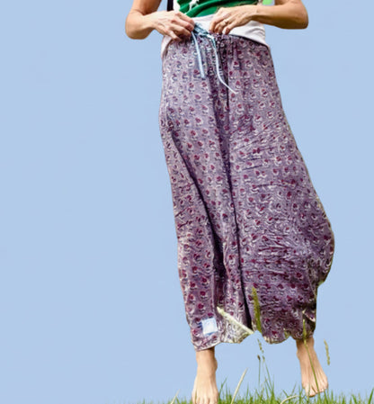 A woman stands outdoors in front of a stone wall and window, wearing loose purple patterned pants and a green-and-white top, with a black crossbody bag across her chest.